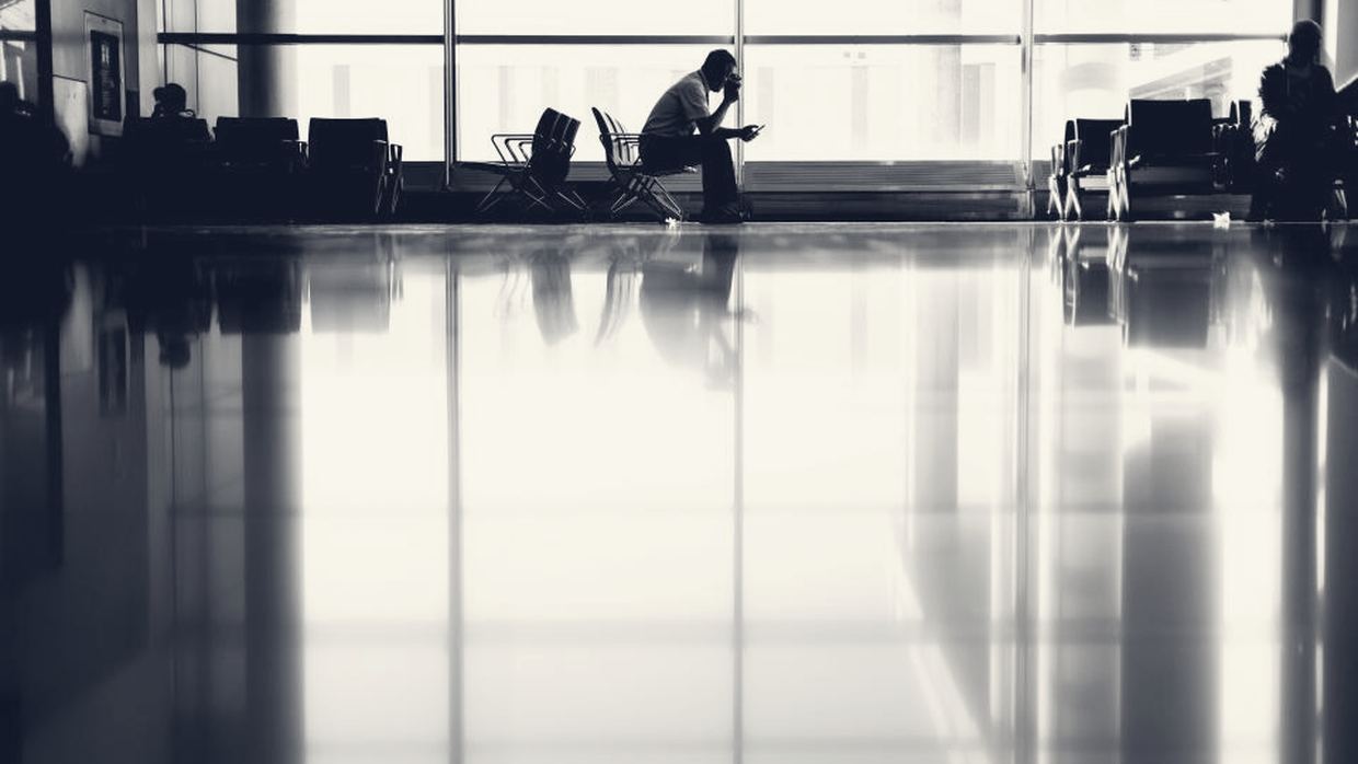 Une personne seule, en contre-jour, assise face aux vitres d'un terminal d'aéroport ; le sol ciré renvoie la lumière.