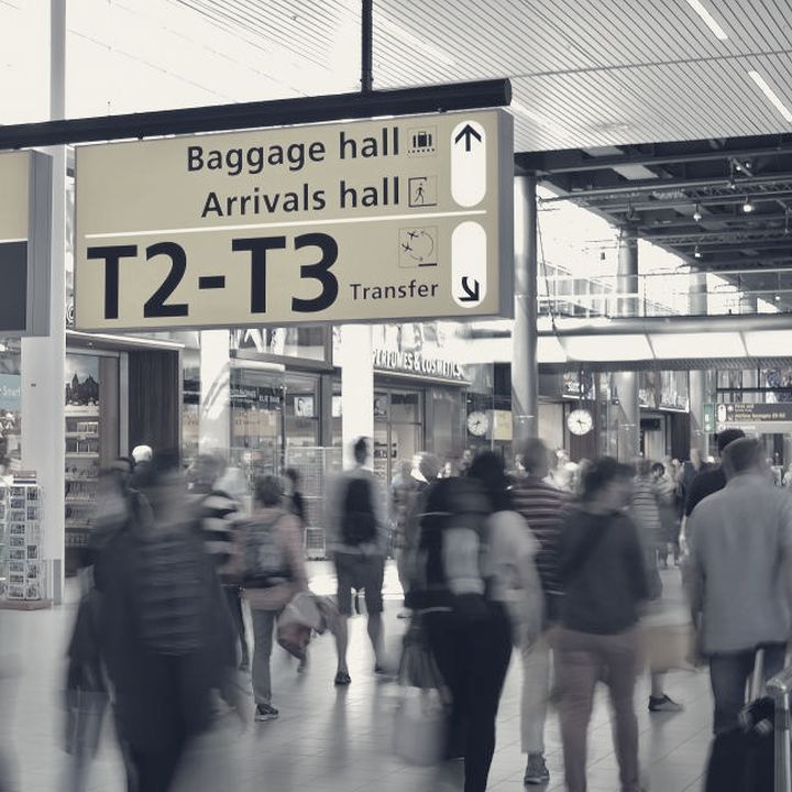 Les panneaux suspendus d'un hall d'aéroport européen indiquent les terminaux et la salle de livraison des bagages, au-dessus d'une foule floue.